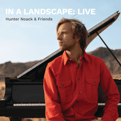 Hunter Noack sits in front of his piano and looks to the side. Mountains in the background.