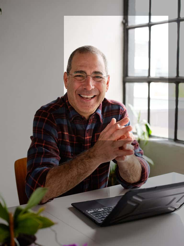 Adam Nemer headshot. Smiling and sitting at a laptop with a window in the background.